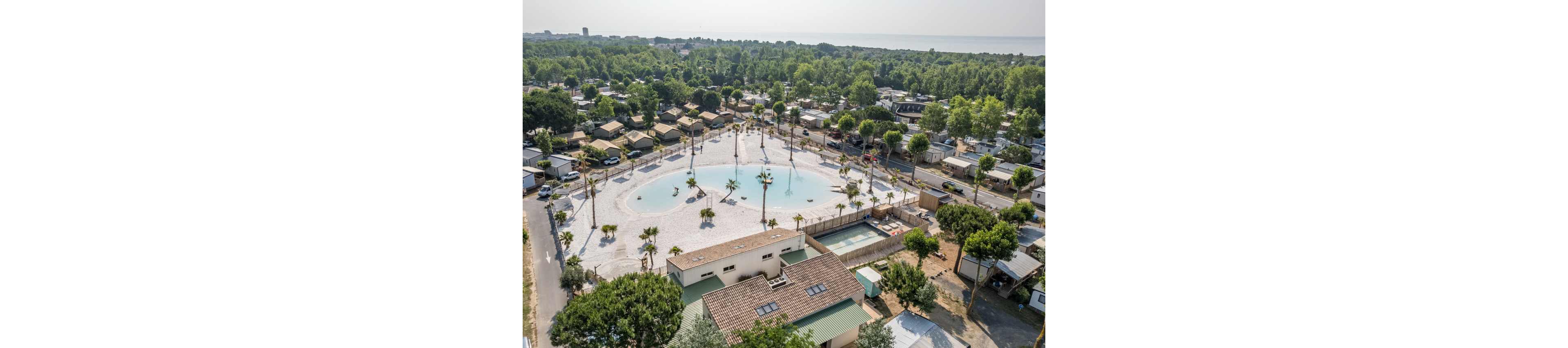 A large holiday resort with a central shallow pool is seen from above. The complex includes numerous cabins and mobile homes arranged among trees and roads. A circular lagoon-style water area with palm trees is surrounded by sunbathing space and nearby rectangular swimming pools. Dense green forest stretches toward the coastline and sea in the background. The overall scene suggests a relaxed vacation atmosphere in a coastal campground setting.
