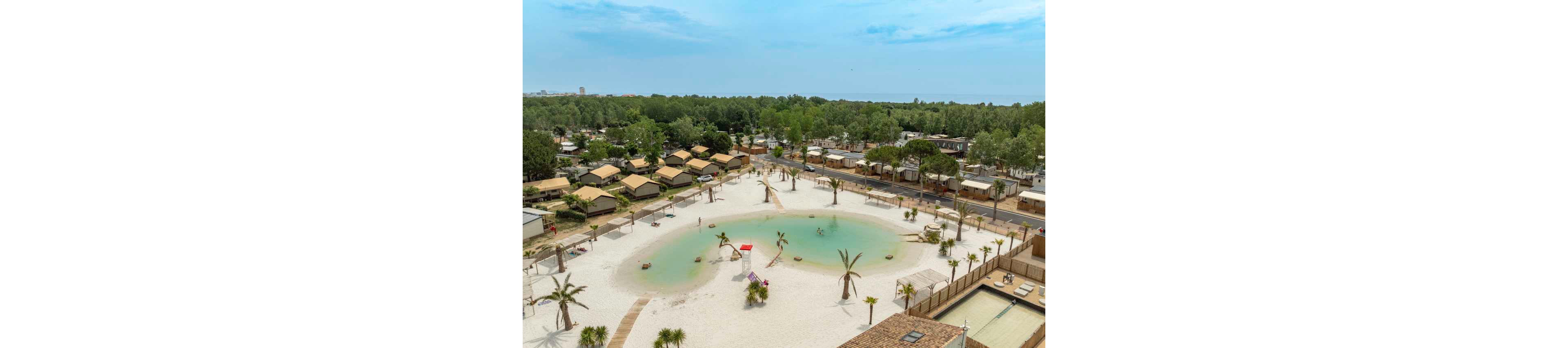 A large artificial lagoon with white sand and scattered palm trees is seen from above in a holiday resort. Wooden cabins and mobile homes surround the lagoon, separated by a road and pathways. The water is shallow and turquoise, with a small lifeguard post and beach umbrellas visible. Dense green forest stretches into the distance under a bright, partly cloudy sky.