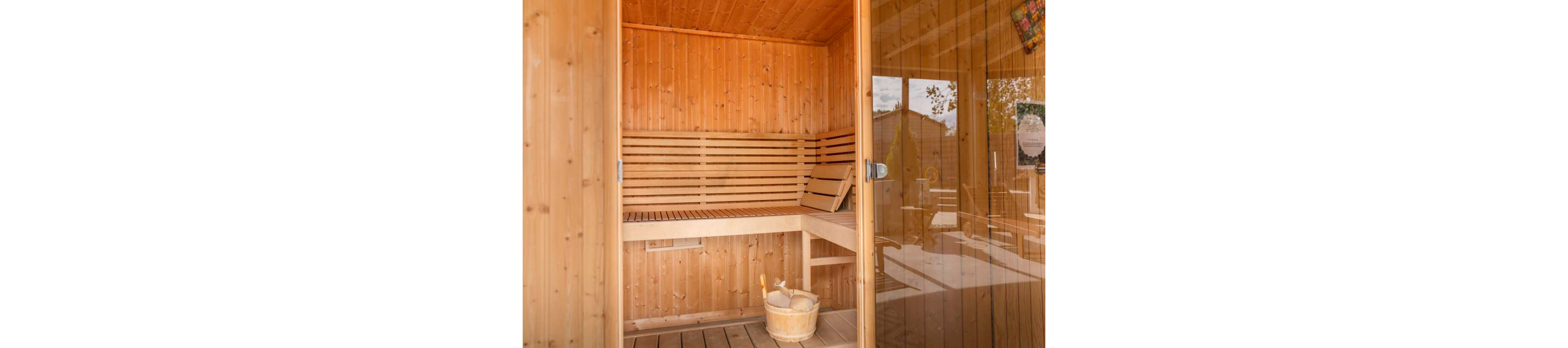 A woman wrapped in a towel reclines on a wooden bench inside a sauna. The interior is entirely lined with warm-toned wood panels and slats. A wooden bucket with a ladle sits on the floor near the bench, emphasizing the spa environment. Sunlight filters through the glass door, creating a calm and peaceful atmosphere.