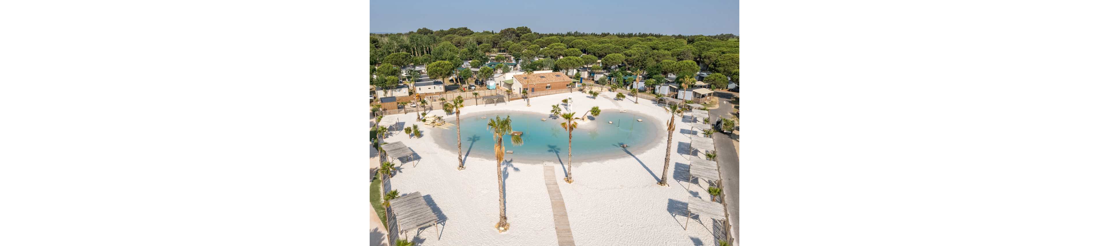 A sandy, man-made lagoon with turquoise water sits at the center of a large campsite. Wooden walkways cross the white sand, leading to shaded cabanas and tall palm trees around the water. Surrounding the lagoon are mobile homes, RVs, and small buildings nestled among dense green trees. The bright daylight and clear sky create a relaxed, vacation atmosphere in this resort-style camping area.