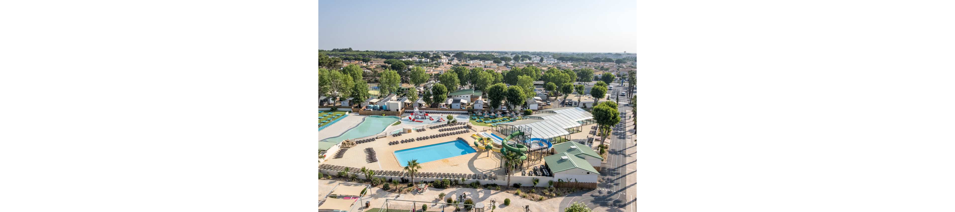 A large outdoor waterpark resort is seen from above, featuring multiple swimming pools and winding water slides. Sun loungers are neatly arranged around the pools, with shaded seating and covered pool areas nearby. The complex is surrounded by trees, small buildings, and a wider residential neighborhood in the background. Clear skies and bright daylight create a relaxed vacation atmosphere.