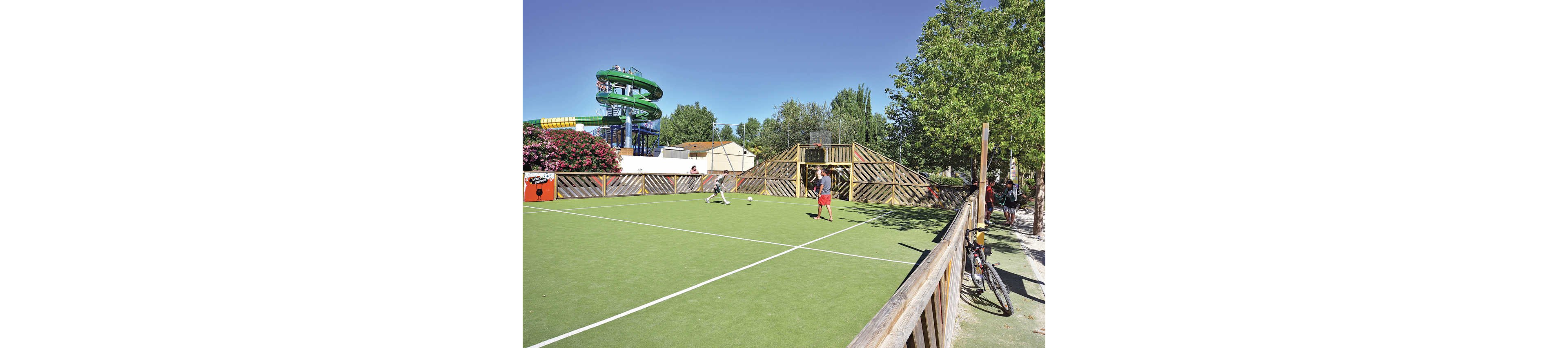 Two children play soccer on a small outdoor sports court with green artificial turf. Wooden fences and a raised viewing platform surround the playing area. In the background, a large green water slide structure rises above trees and nearby buildings, suggesting a holiday park or campground. Bicycles are parked along the fence, and the scene is bright and sunny with clear blue skies.