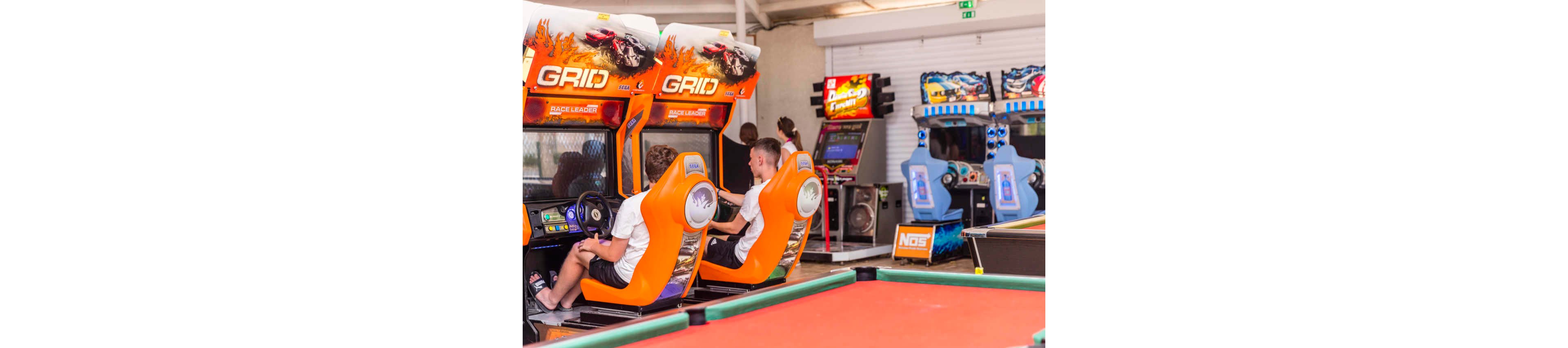 Two teenage boys sit in bright orange GRID racing arcade machines in a lively game room. The players face large screens with racing graphics while gripping the steering wheels. Additional arcade cabinets and machines are visible in the background, adding depth and color to the scene. The overall atmosphere is energetic and fun, with a focus on competitive gaming and social entertainment.