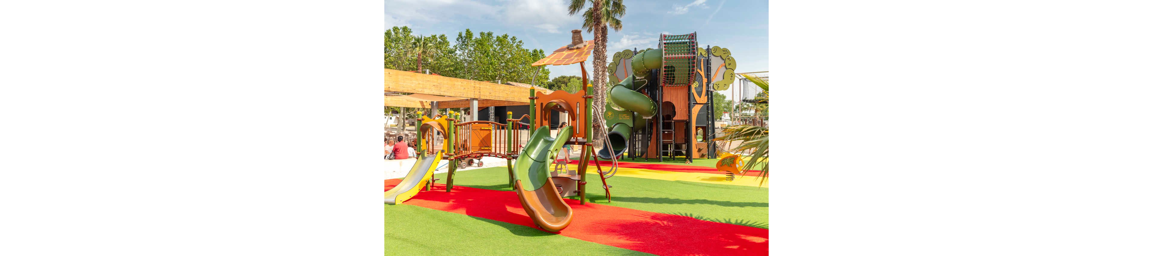 A vibrant outdoor playground features multiple slides, climbing structures, and towers in green, orange, and brown tones. The play area is set on bright artificial turf with red and green sections, creating a lively and safe surface. Palm trees and leafy trees surround the playground, suggesting a warm, sunny climate. A few people sit in the background while the equipment stands ready for children to play.