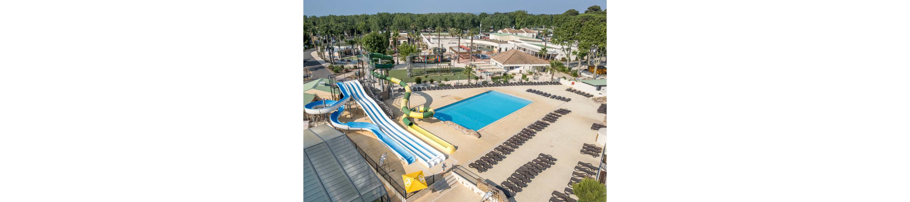 A large outdoor water park features multiple colorful water slides leading into a pool. The central rectangular swimming pool is surrounded by rows of empty lounge chairs on a spacious deck. Nearby buildings, trees, and additional recreational areas indicate a resort or campground setting. The bright daylight and clear sky create a sunny, inviting atmosphere for vacation and leisure.
