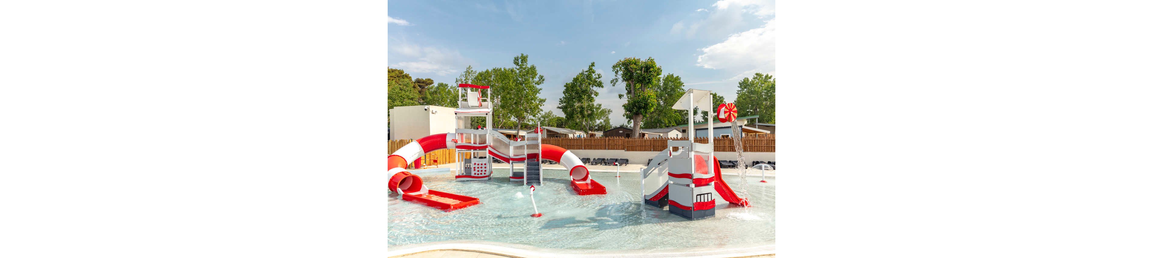 An outdoor water playground features bright red and white slides and climbing structures in a shallow pool. Multiple water sprayers and fountains create playful splashes around the equipment. The area is surrounded by a wooden fence, trees, and nearby buildings under a clear blue sky. The scene suggests a family-friendly aquatic park designed for children.