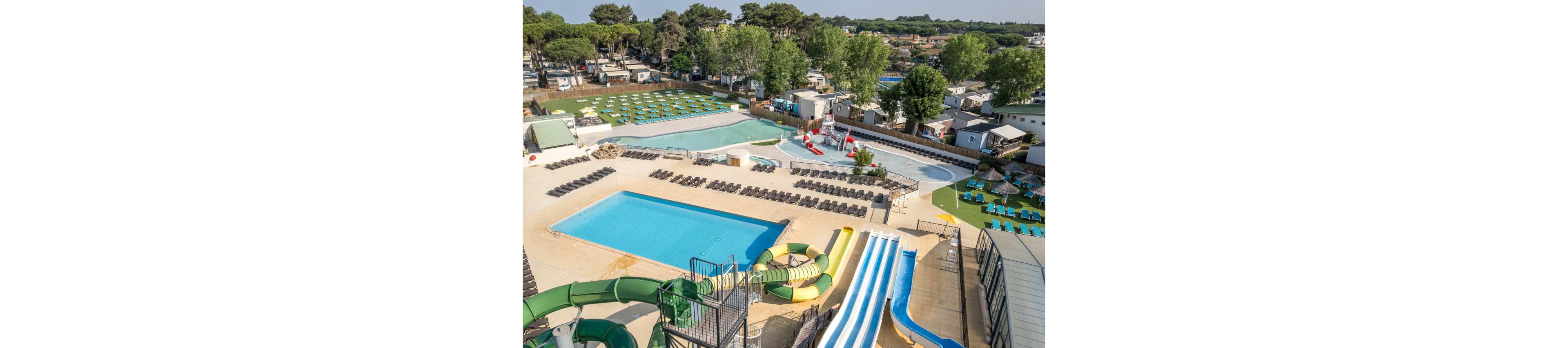 A large outdoor water park complex is shown from above, featuring multiple swimming pools and colorful water slides. Rows of sun loungers surround the pools, indicating a leisure and vacation setting. Nearby, mobile homes and cabins suggest the water park is part of a holiday resort or campground. The scene is bright and sunny, with green trees and landscaped areas adding to the relaxed, family-friendly atmosphere.
