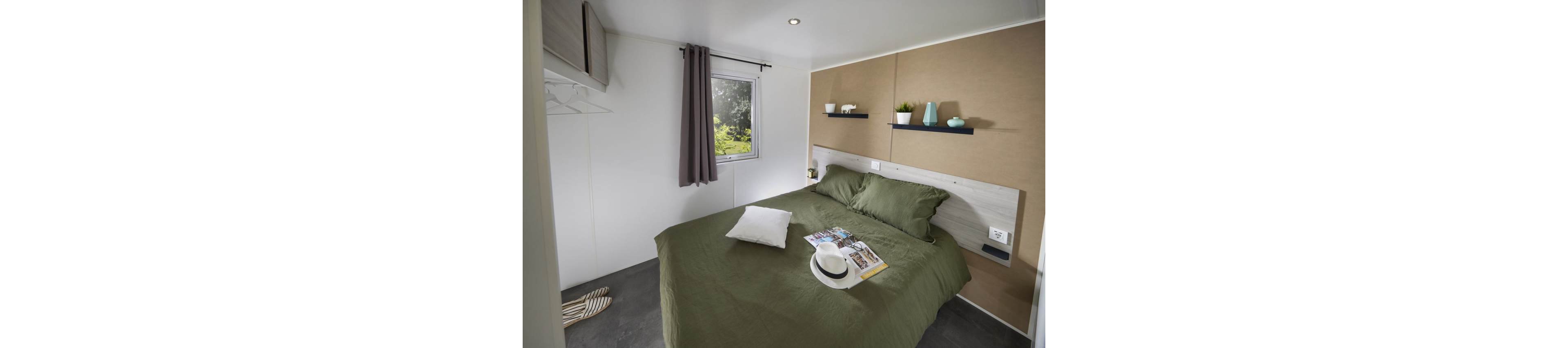 A compact modern bedroom features a double bed with green bedding and a single white pillow. A magazine, sunhat, and sunglasses rest on the bed, suggesting leisure or vacation. The room includes a small window with a dark curtain, wall shelves holding decorative plants and vases, and built-in storage on the left. Neutral walls and soft lighting create a calm, minimalist atmosphere.