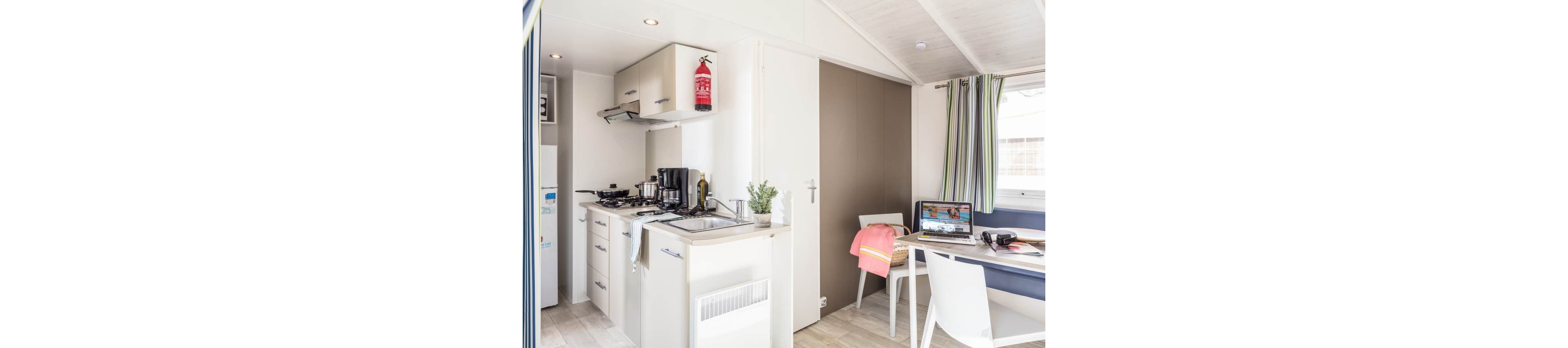 Bright compact studio interior featuring a small kitchen and dining area. The kitchenette includes a gas stove, sink, coffee maker, and overhead cabinets, with a fire extinguisher mounted above. A table beside the bench seating holds an open laptop, camera, and headphones, suggesting a casual work or leisure setup. Light wood flooring, white walls, and striped curtains create a clean, modern holiday home atmosphere.
