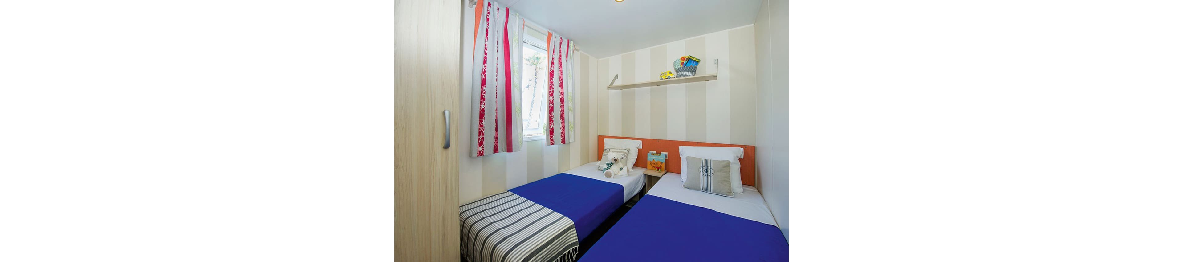 Compact bedroom featuring two single beds with blue bedspreads and striped blankets. The room has light vertical striped walls and a small window with colorful patterned curtains. A shelf above the beds holds decorative items and storage baskets, adding a cozy touch. Overall, the space appears clean, bright, and suitable for guests or children.