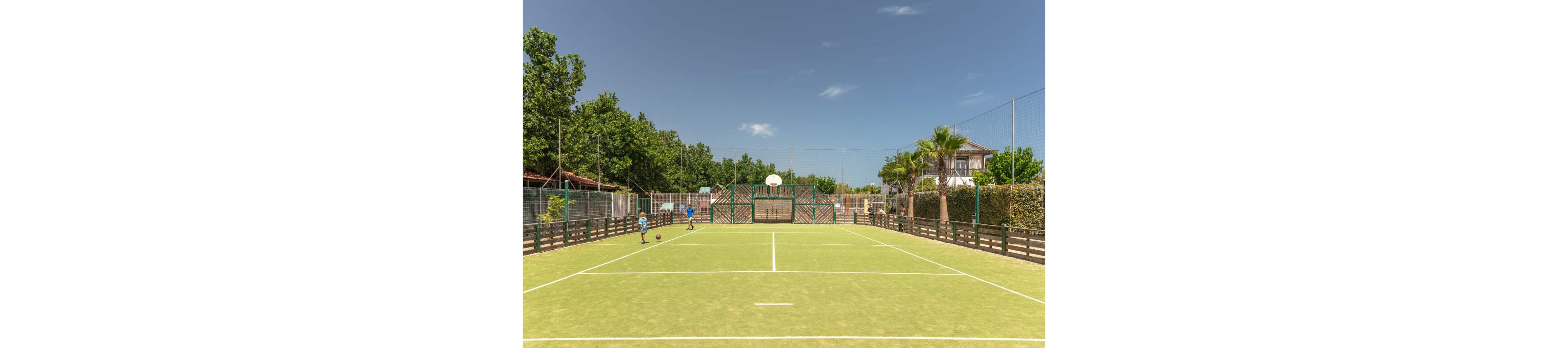 Two children play with a ball on a multi-use outdoor sports court under a clear blue sky. The court features tennis lines, basketball hoops, and high protective fencing around the perimeter. Trees, hedges, and a few residential-style buildings surround the court, creating a relaxed recreational setting. The bright daylight and green surface give the scene a warm, summery atmosphere.