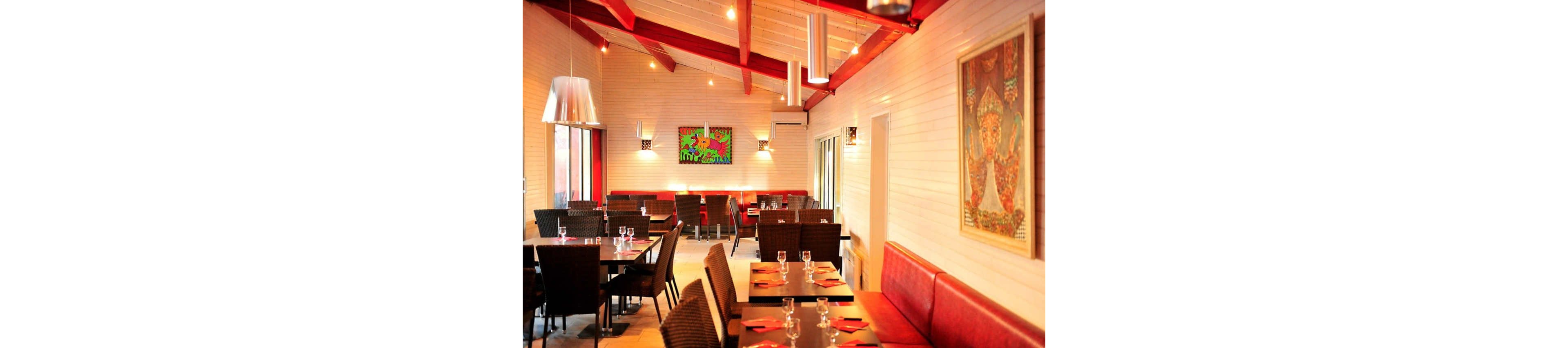 A bright, modern restaurant dining room features neatly arranged tables with glassware and red place settings. Warm pendant lights hang from a high wooden ceiling with exposed beams, casting a soft glow across the space. Wicker-style chairs and a long red banquette line the walls, creating an inviting atmosphere. Colorful artwork decorates the light-toned walls, adding a contemporary touch to the cozy interior.