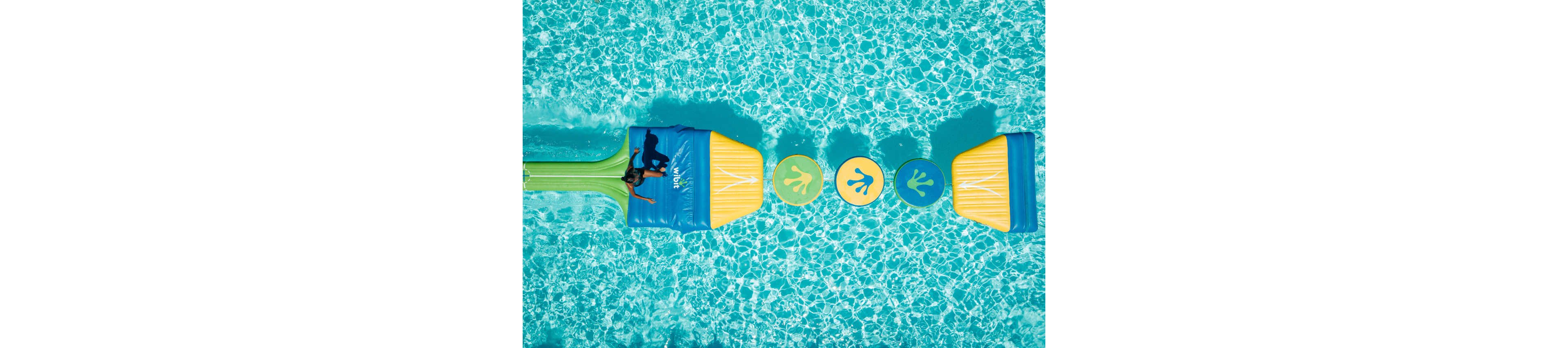 A child plays on a colorful inflatable obstacle course floating in a bright blue swimming pool. The structure features large yellow and green platforms connected by circular pads with footprint graphics. Sunlight reflects off the clear water, creating a sparkling pattern around the floating course. The scene feels playful and summery, emphasizing outdoor fun and recreation.
