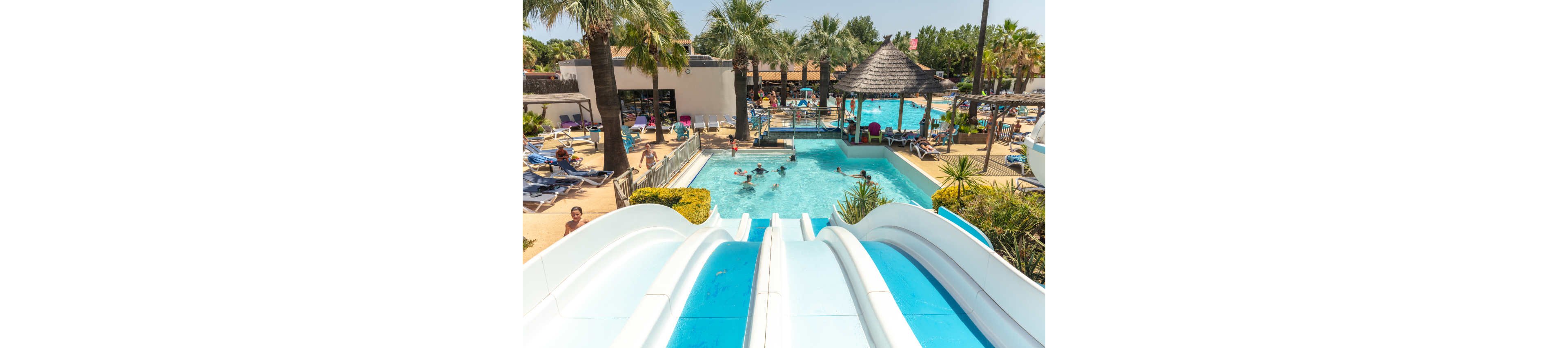 A bright resort swimming pool features multiple white water slides leading into the clear blue water. Children and adults play and swim in the pool, surrounded by sun loungers and parasols. Tall palm trees and tropical plants create a vacation atmosphere under a clear sky. The scene suggests a lively family-friendly water park in warm, sunny weather.