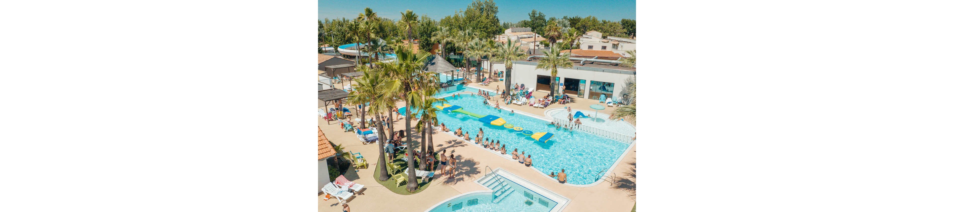 An aerial view shows a busy resort swimming pool surrounded by palm trees and sun loungers. Many people relax on deck chairs while others swim or sit along the pool edge. A floating inflatable obstacle course stretches across the main pool, attracting groups of children and adults. In the background, additional pools, water slides, and low-rise vacation buildings complete the holiday setting.