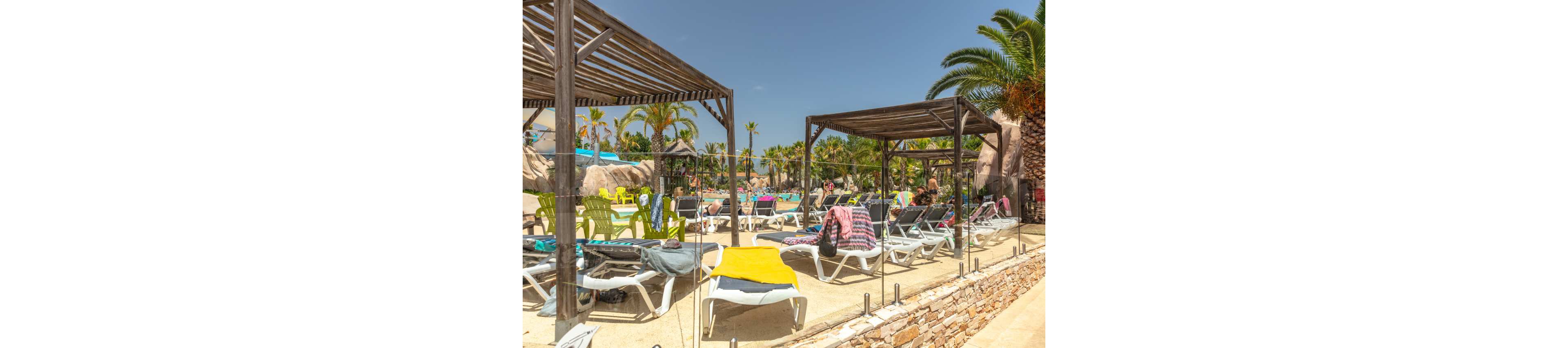 Outdoor resort pool area with numerous sun loungers arranged under wooden pergolas. Colorful towels and bags are draped over the chairs, suggesting guests relaxing nearby. Palm trees, tropical plants, and rock features surround the pool, creating a vacation atmosphere. Bright sunlight and clear blue sky emphasize a warm, summery day at the resort.