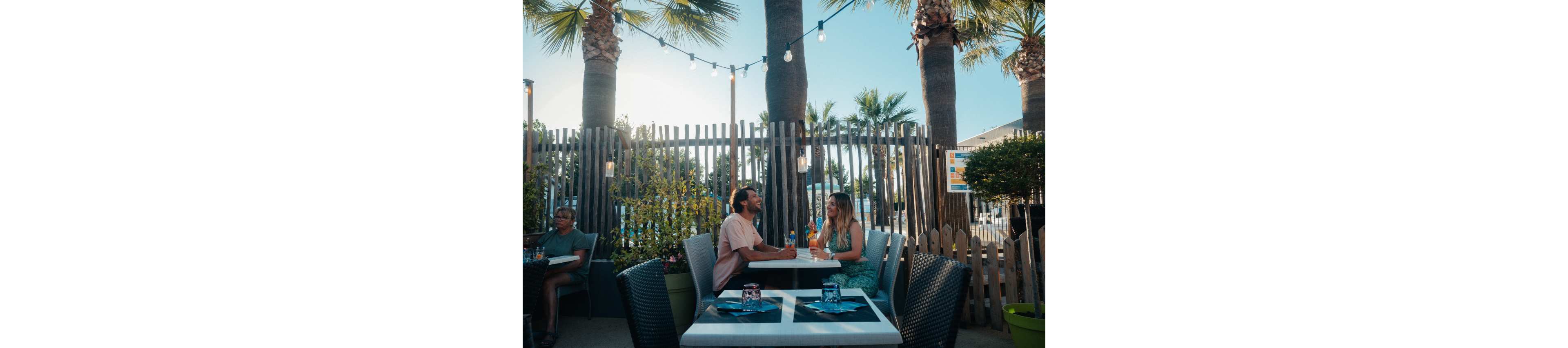 A young couple sits across from each other at an outdoor restaurant patio, holding colorful drinks and talking. The setting features palm trees, string lights, and wooden fencing that create a relaxed, summery atmosphere. Other diners are visible in the background, adding to the casual social scene. Warm sunlight and soft shadows enhance the vacation-like mood of the gathering.