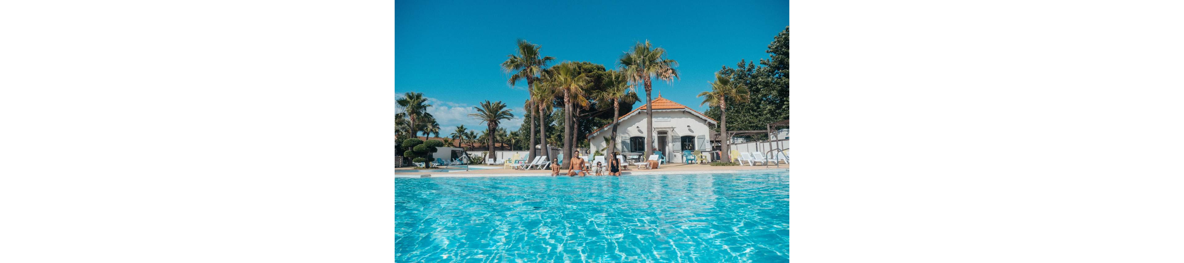 A family sits at the edge of a large outdoor swimming pool under a clear blue sky. Palm trees surround the pool area, creating a tropical resort atmosphere. A white villa-style building with an orange roof and sun loungers is visible in the background. The bright sunlight and turquoise water give the scene a warm, summery vacation feel.
