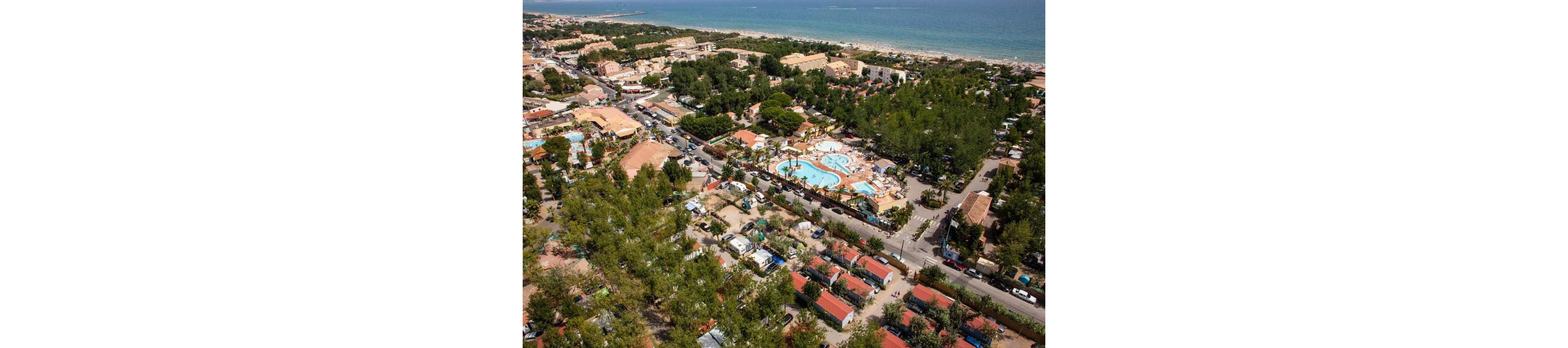 A large holiday resort with multiple swimming pools is captured from an aerial perspective near a coastline. The complex includes red-roofed cabins, campsites, and tree-lined roads surrounded by dense greenery. People can be seen enjoying the pools and outdoor facilities under clear weather. In the background, a sandy beach and calm sea stretch along the horizon, emphasizing the seaside vacation setting.