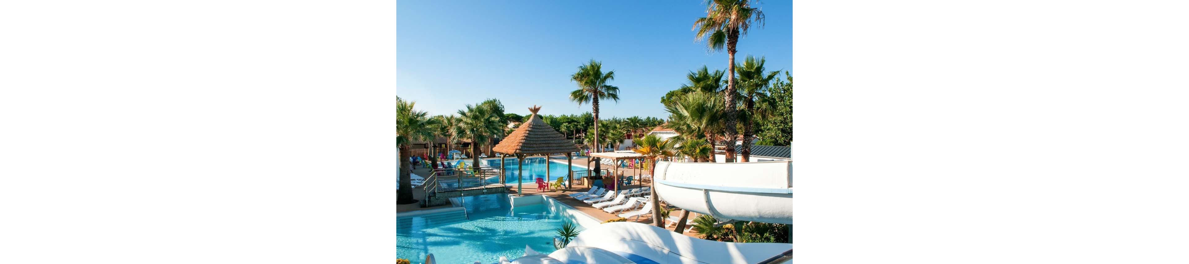A large outdoor resort pool is surrounded by palm trees and sun loungers under a clear blue sky. A white waterslide curves into the pool area, adding a sense of fun and activity. Thatched-roof structures and shaded seating areas create a tropical, vacation-like atmosphere. The scene suggests a relaxing holiday destination with bright, summery colors and inviting water.