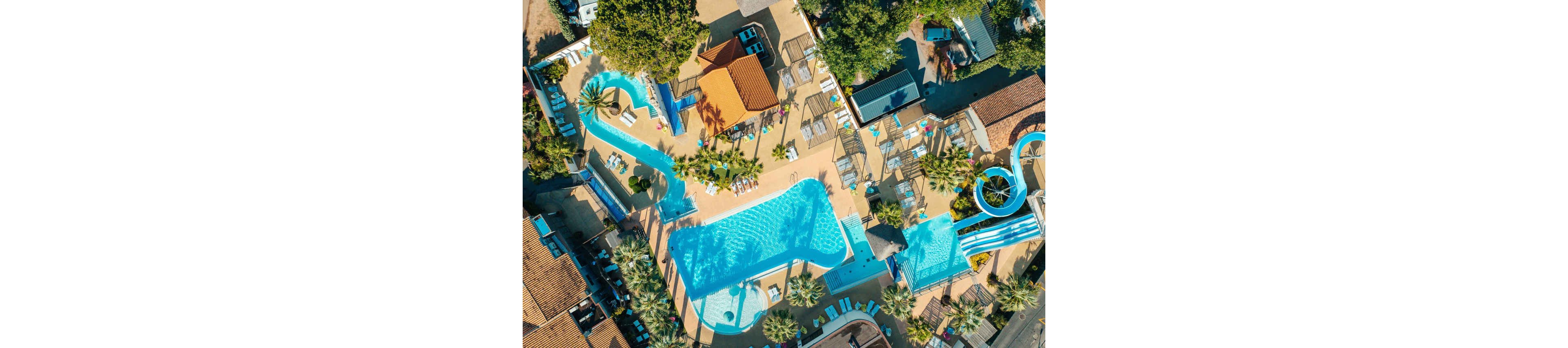 A large outdoor swimming pool complex is captured from above in bright daylight. The scene includes multiple pools, winding water slides, and sun loungers arranged around the deck. Buildings with terracotta roofs and surrounding trees frame the aquatic area, creating a vacation resort atmosphere. The turquoise water contrasts with the warm tones of the paving and rooftops, suggesting a sunny, relaxing holiday setting.
