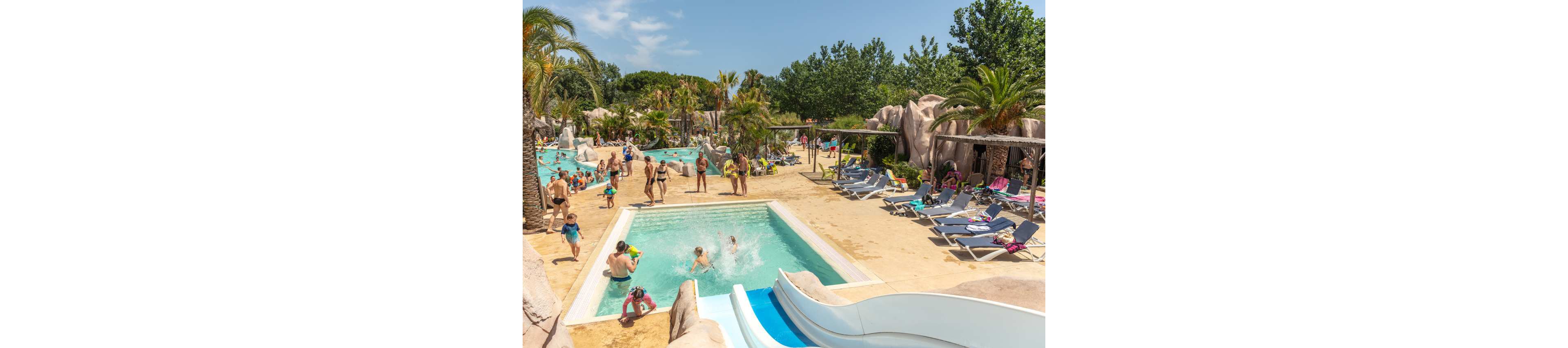 A lively outdoor swimming pool area is filled with families enjoying a sunny day. Children play in the shallow pool and slide down a white water slide while adults relax nearby. Palm trees, rocks, and greenery create a tropical resort atmosphere around the pools. Sun loungers and parasols line the deck, suggesting a vacation setting with warm weather and leisure activities.