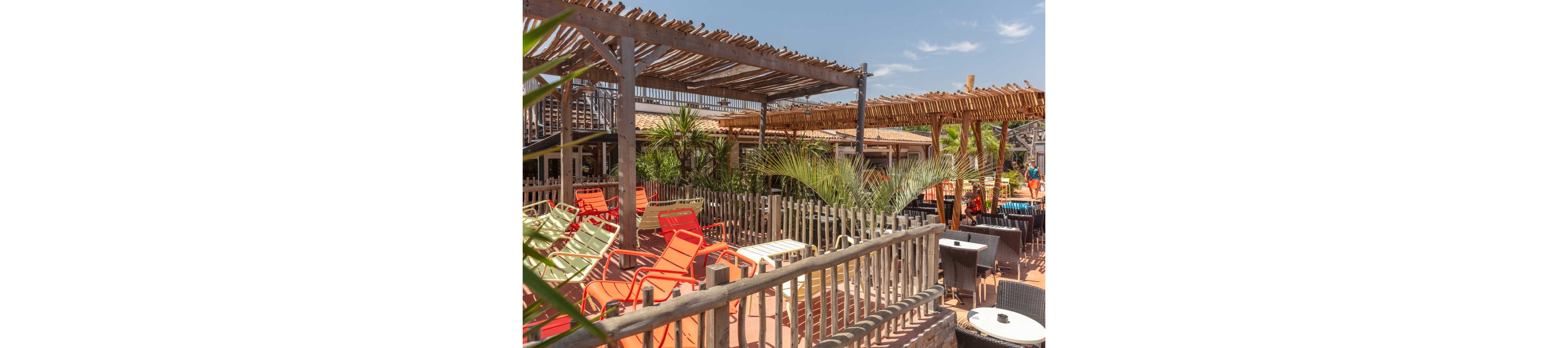 An outdoor beach bar terrace features colorful lounge chairs arranged under wooden pergolas. The setting includes rustic wooden railings, potted palms, and tropical plants creating a relaxed vacation atmosphere. Bright sunlight and clear blue sky suggest a warm summer day. Empty seating areas and sun loungers indicate a tranquil moment before guests arrive.