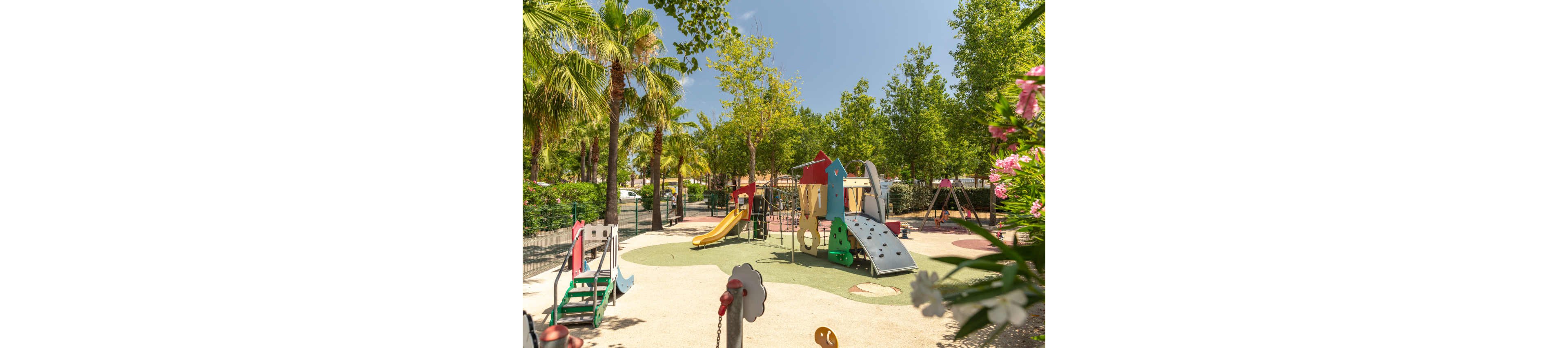 A bright outdoor playground features colorful slides, climbing structures, and swings surrounded by lush green trees. The play area is set on a soft, light-colored surface under clear blue skies. Palm trees and flowering plants frame the scene, creating a warm and inviting atmosphere. No people are present, emphasizing the playground equipment and tranquil park setting.