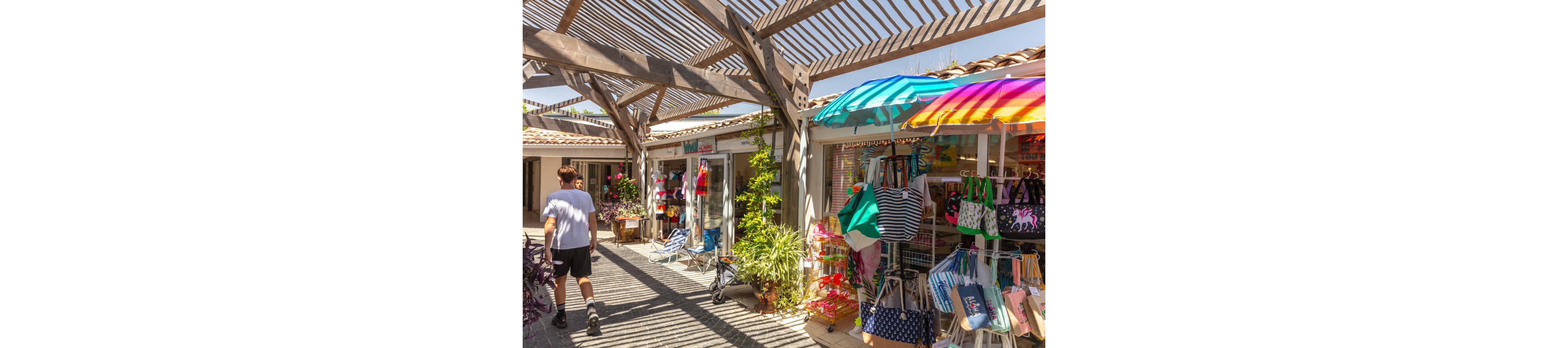 An outdoor market corridor displays colorful beach bags, towels, and summer accessories under striped umbrellas. Wooden pergolas cast patterned shadows on the walkway, creating a bright and sunny atmosphere. A person walks past small shops filled with clothing, hats, and souvenirs. Potted plants and flowers add greenery and charm to the relaxed shopping scene.