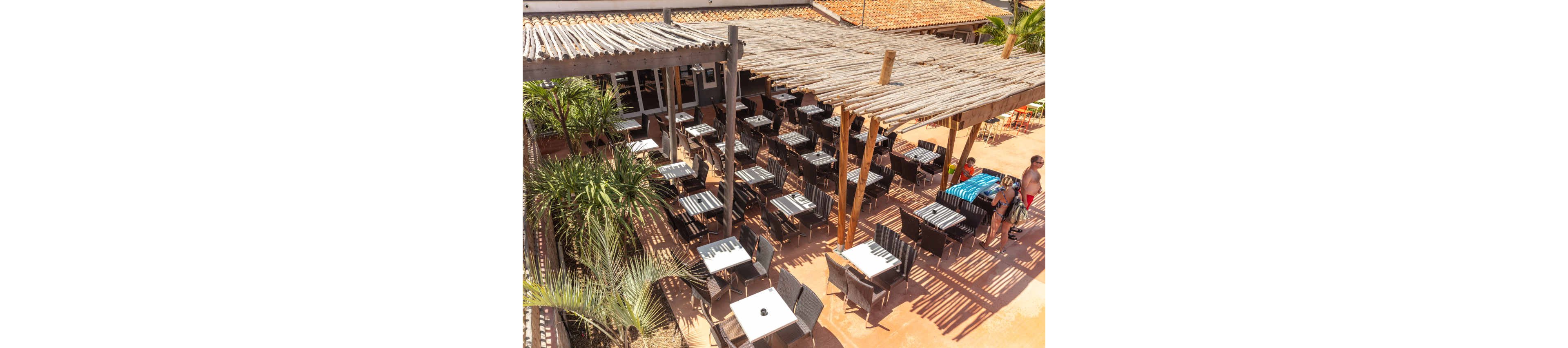 An elevated view shows a spacious outdoor café terrace with rows of empty tables and chairs neatly arranged. Wooden pergolas provide partial shade over the seating area, casting striped shadows on the ground. Lush green palm plants frame the scene, adding a tropical touch. A few casually dressed people stand near the edge of the terrace, suggesting a relaxed holiday atmosphere.