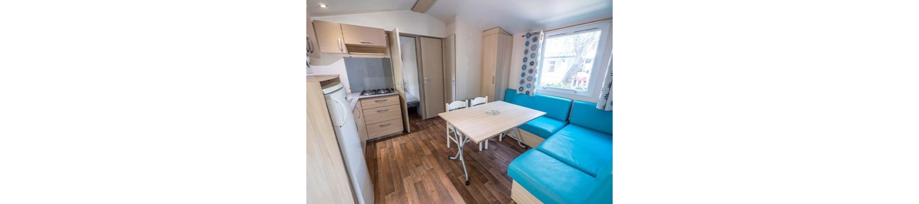 Bright interior of a compact mobile home living space with a small kitchenette and dining area. A light wooden table with white folding chairs stands beside a turquoise corner sofa. The kitchenette includes cabinets, a gas cooktop, and a small refrigerator, with a doorway leading to a bedroom. Large windows with patterned curtains let in natural light, highlighting the wood-style flooring and cozy, functional layout.