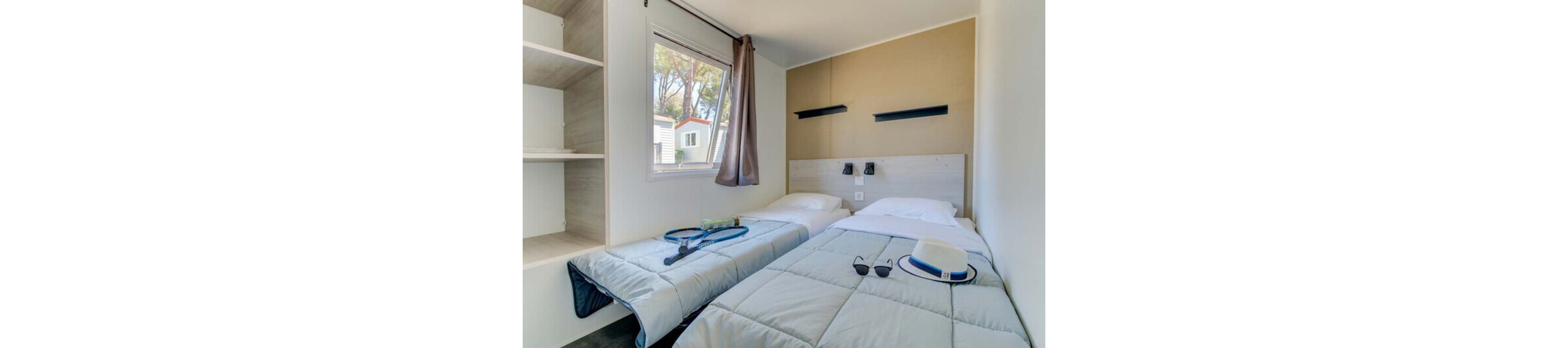 A compact bedroom features two neatly made twin beds with light gray comforters. The room is bright, with daylight coming through a window that shows neighboring cabins and tall trees outside. A tennis racket and ball rest on one bed, while a hat and sunglasses lie on the other, suggesting a relaxed holiday stay. Built-in shelving, wall-mounted reading lights, and neutral decor create a clean, modern atmosphere.