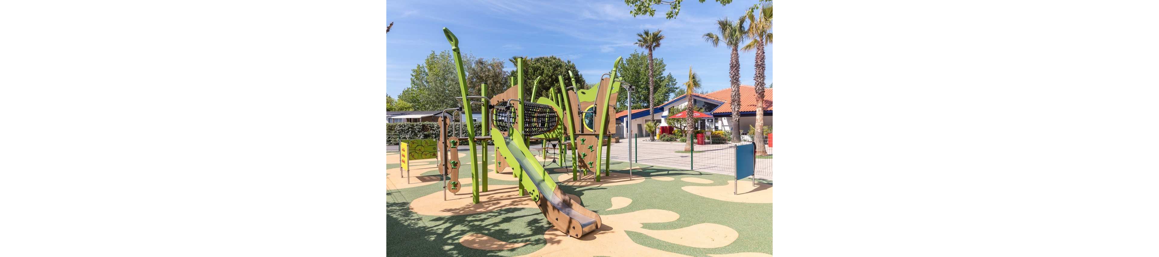 A colorful modern playground features green and beige climbing structures and a slide in an outdoor recreational area. The play equipment includes tunnels, ladders, and elevated platforms set on a patterned rubber surface. Palm trees and other greenery surround the space, while low buildings and a fenced perimeter define the background. The scene is bright and sunny, suggesting a warm, family-friendly environment.