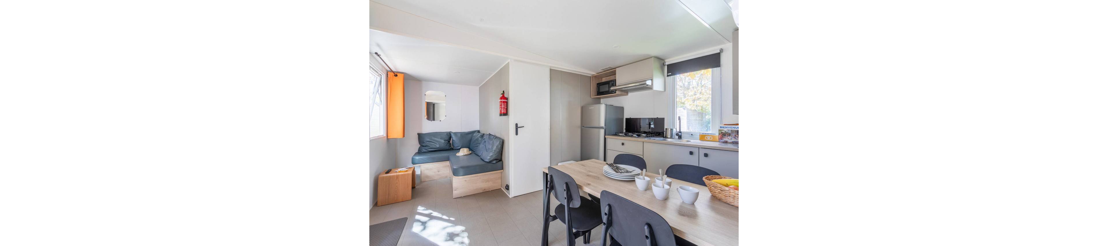 Interior view of a compact mobile home featuring a combined kitchen and dining area. A wooden dining table with black chairs is set with plates, bowls, and a wicker basket. The kitchenette includes a refrigerator, stovetop, microwave, and cabinets along one wall. A small lounge area with a blue cushioned bench and window with orange curtain is visible in the background. Natural light from the windows brightens the clean, modern space.
