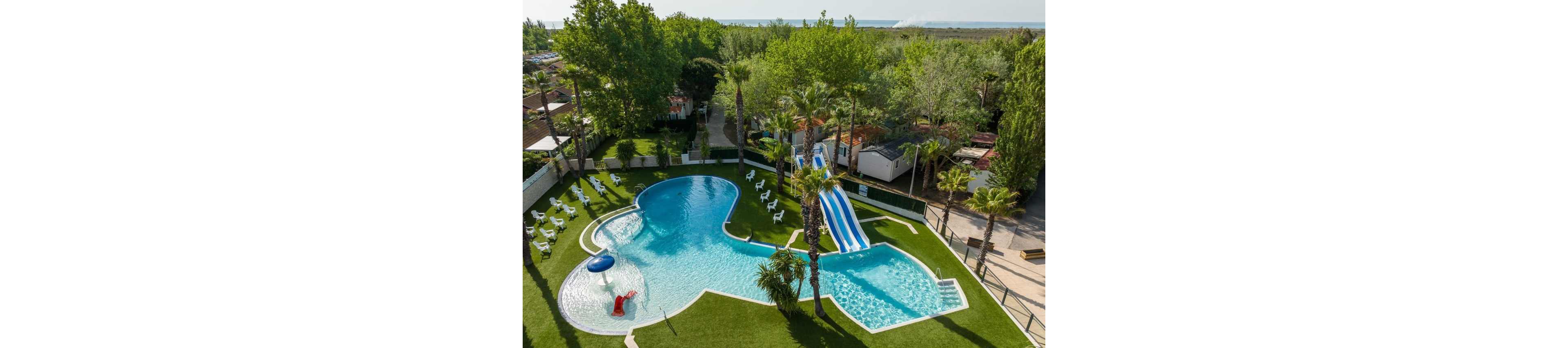 An aerial view shows a resort-style swimming pool complex surrounded by lush green lawns and tall trees. The uniquely shaped pool includes a shallow area with a small red slide and a deeper section connected to multiple blue-and-white water slides. White lounge chairs are neatly arranged around the pool, creating a relaxing sunbathing area. Nearby buildings and pathways suggest a holiday park or campground setting close to nature.