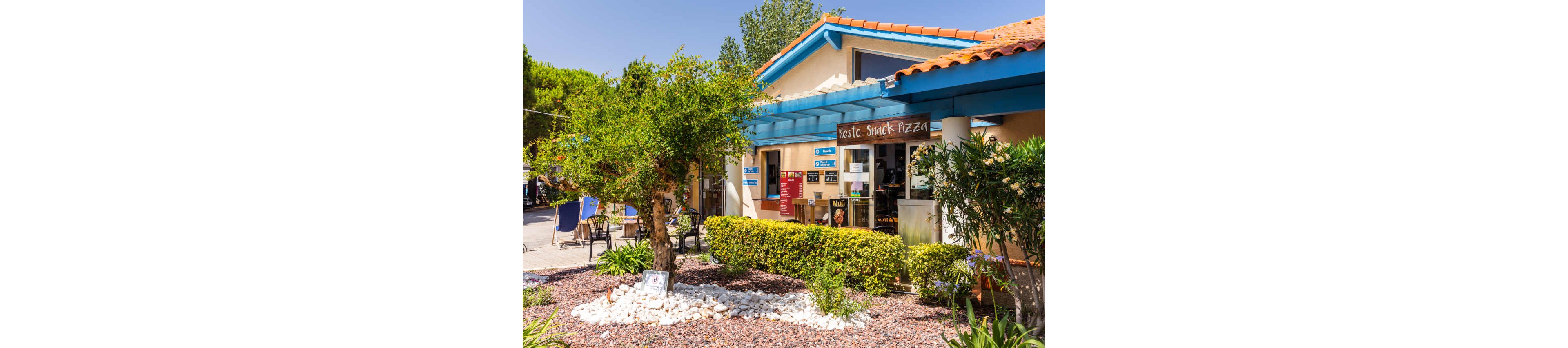 A small snack bar and pizza restaurant is shown in a sunny outdoor setting. The building has a tiled roof, blue beams, and a shaded entrance with various signs and menus. In the foreground, a landscaped garden with a tree, white stones, and shrubs decorates the front of the establishment. Patio chairs and greenery suggest a relaxed, casual atmosphere for visitors. The scene conveys a warm, inviting place to eat and rest.