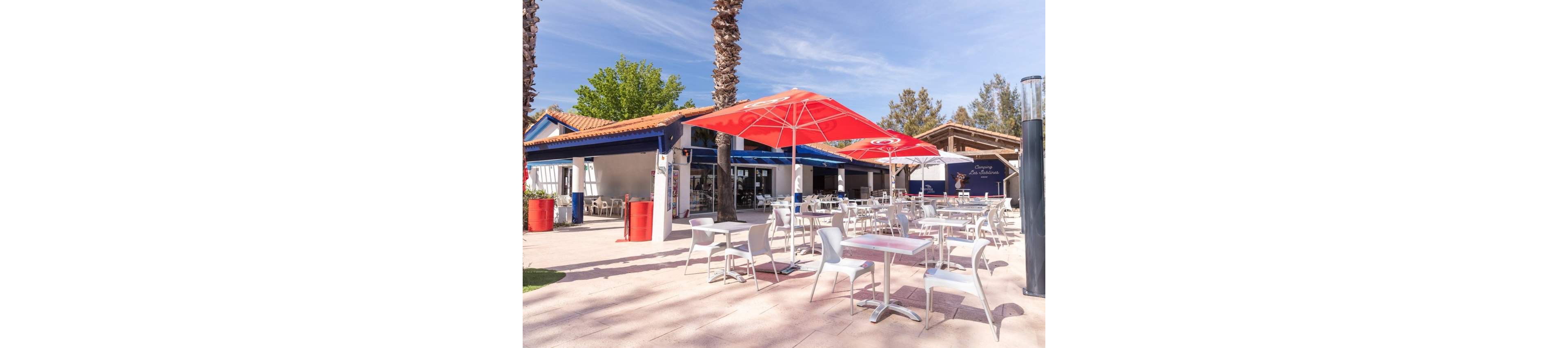 An open-air café terrace features multiple white tables and chairs arranged neatly under large red umbrellas. The setting appears to be a sunny day with clear blue skies and tall palm trees providing additional shade. A low building with a tiled roof and blue-and-white façade forms the backdrop, with open doors suggesting a casual dining area inside. The scene is quiet and empty, emphasizing the relaxed, inviting atmosphere of the outdoor seating area.