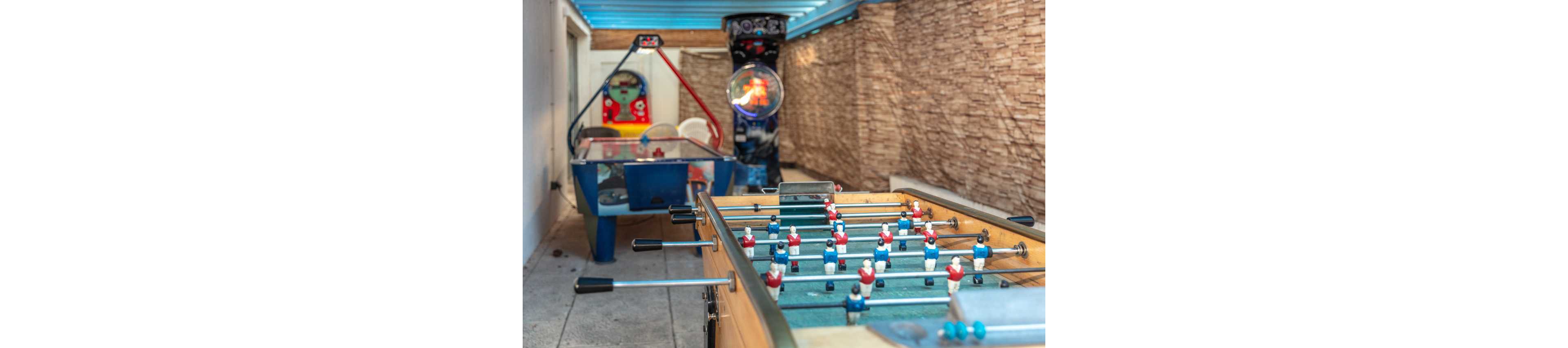 A foosball table with red and white players stands in the foreground of an indoor game room. Behind it, an air hockey table and an upright arcade machine line the narrow space. The room features stone-textured walls and a light blue ceiling structure, creating a casual entertainment atmosphere. The perspective draws the viewer’s eye down the row of games toward the back wall.