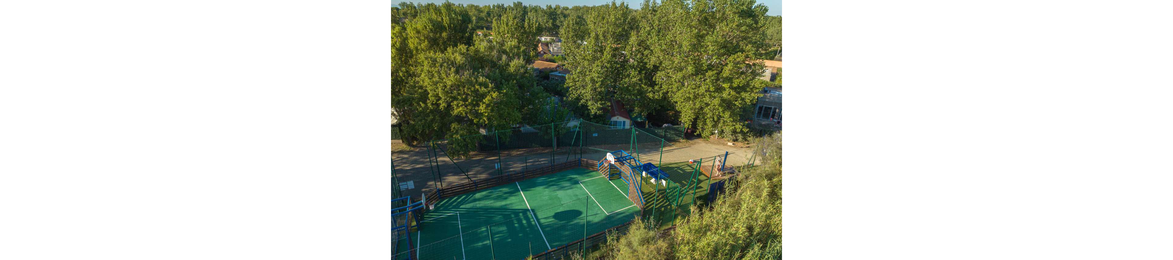 An aerial view shows an outdoor sports court surrounded by tall green trees. The fenced court features a green playing surface with white boundary lines and a small goal structure at one end. Nearby, camping structures and buildings are partially visible among the trees. The scene suggests a recreational area within a campsite or holiday park on a sunny day.