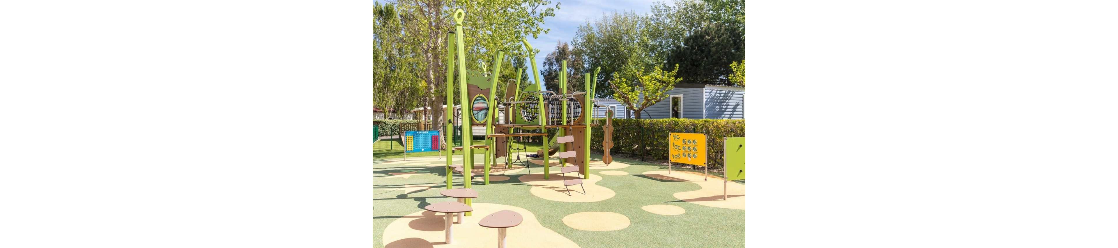 An empty modern playground with green and beige equipment stands in a sunny outdoor area. The play structure includes climbing nets, platforms, and stepping pods arranged on a soft rubber surface with circular patterns. Brightly colored activity panels and trees surround the play area, while mobile homes or cabins are visible in the background. The scene suggests a quiet, family-friendly campsite or holiday park on a clear day.