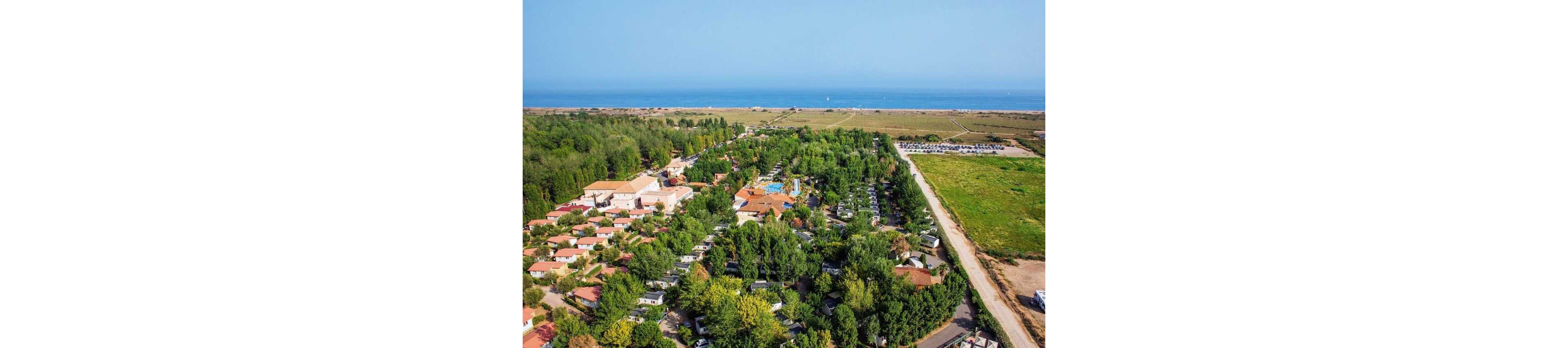An aerial view shows a large holiday resort surrounded by dense green trees near the coastline. Rows of small houses and cabins are arranged along narrow internal roads, with a central complex that appears to include a pool area. Open fields and a long straight road border the resort on one side, while the sea and sandy shoreline stretch across the horizon. The clear sky and calm water suggest a warm, pleasant day at a coastal vacation destination.