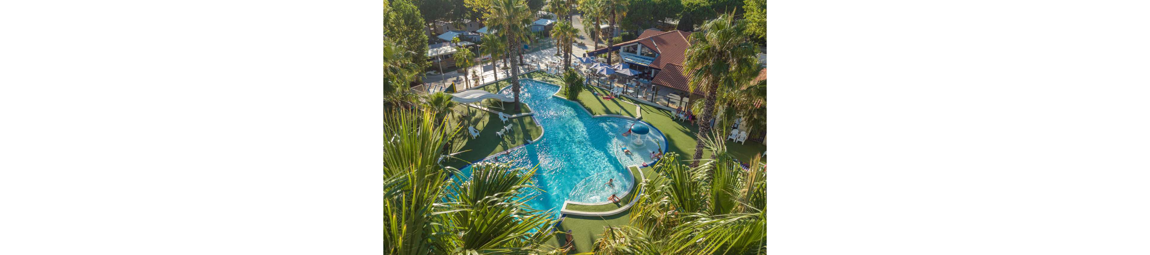 An aerial view shows a large, curving resort swimming pool surrounded by lush palm trees. People relax and play in the clear blue water, with some lounging on chairs along the artificial grass deck. Nearby buildings and outdoor seating areas suggest a vacation campground or holiday resort setting. The bright sunlight and greenery create a tropical, family-friendly atmosphere.