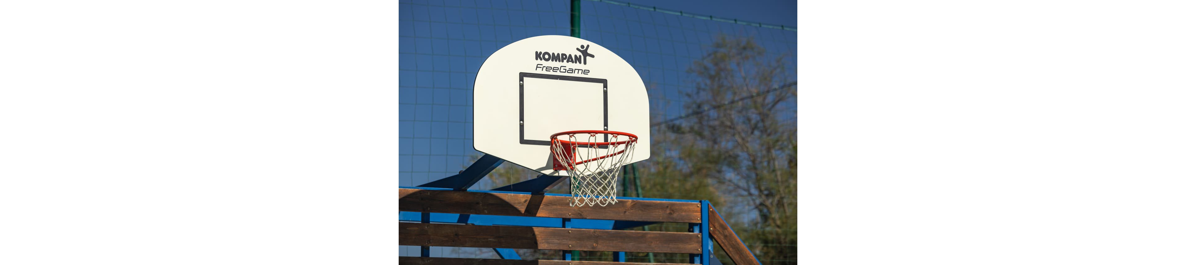 An outdoor Kompan FreeGame basketball hoop is mounted on a wooden and metal structure. The white backboard and metal hoop stand out against a clear blue sky and a green netted fence in the background. Trees with early foliage are visible behind the court, suggesting a park or recreational area. The scene conveys a casual, community sports environment in bright daylight.