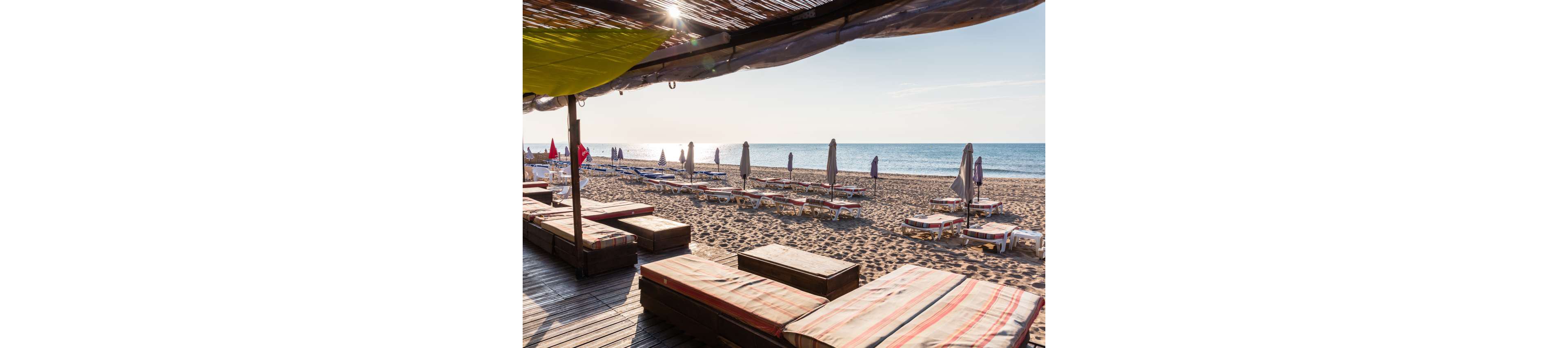 A tranquil beach resort features rows of sun loungers and closed umbrellas facing the calm sea. Wooden deck platforms with striped cushions sit in the foreground under a shaded canopy. The sandy shoreline stretches into the distance under a clear sky. The scene suggests an early morning or off-season moment before guests arrive.