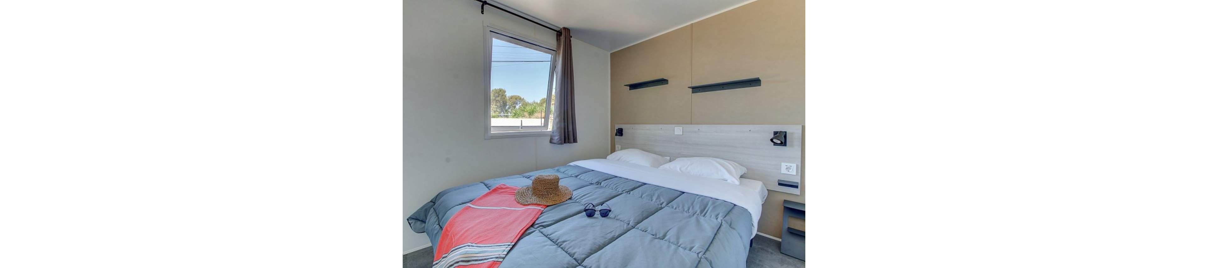 A neatly made double bed with a blue comforter is the main focus of this modern bedroom. The room features a large window with a sheer curtain, allowing natural light to brighten the neutral-toned walls. A straw hat and sunglasses rest on a coral and gray towel at the foot of the bed, suggesting a relaxed vacation atmosphere. Minimalist wall shelves and bedside lighting add to the clean, contemporary design.