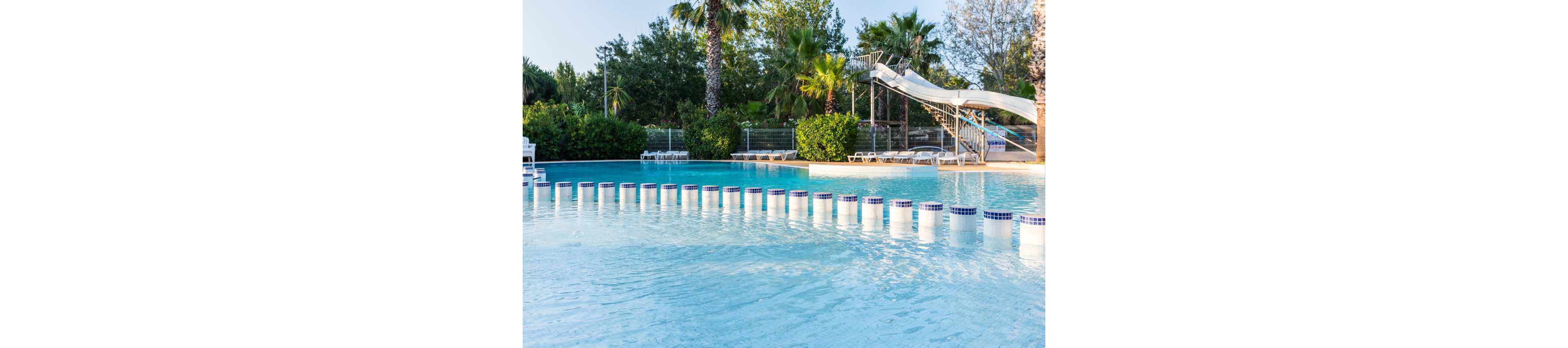 An outdoor swimming pool features a row of cylindrical stepping stones across shallow water. In the background, a tall metal waterslide structure leads down into the pool area. Palm trees and lush green shrubs surround the pool, creating a tropical resort atmosphere. White lounge chairs line the deck behind a fence, suggesting a relaxing leisure setting. The scene is brightly lit, indicating a sunny day.
