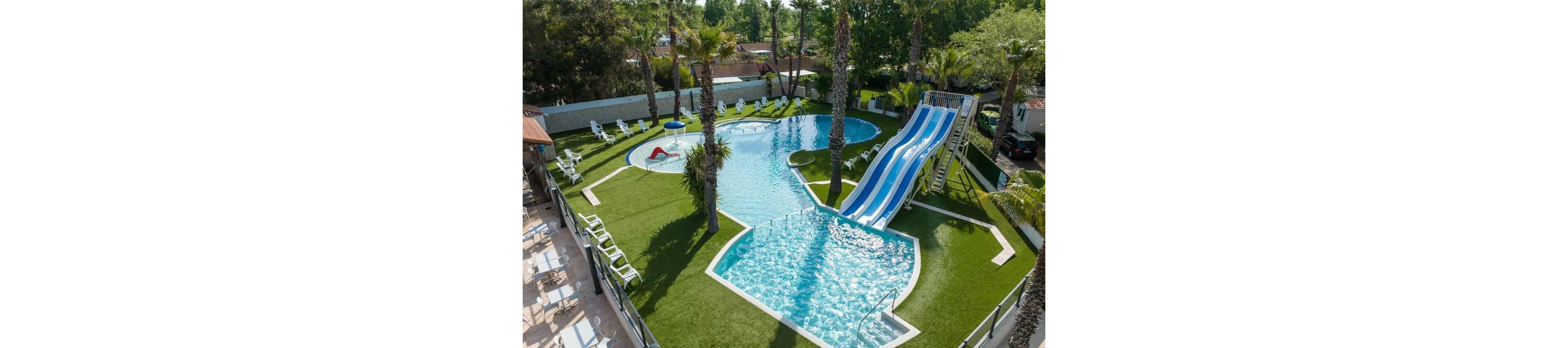 An aerial view shows an outdoor resort-style swimming pool with curving shapes and clear blue water. A large multi-lane water slide descends into one section of the pool, while another shallow area is separated by a railing. Artificial grass surrounds the pool, lined with white lounge chairs and palm trees providing a tropical atmosphere. The setting appears to be a family-friendly recreational area on a sunny day.