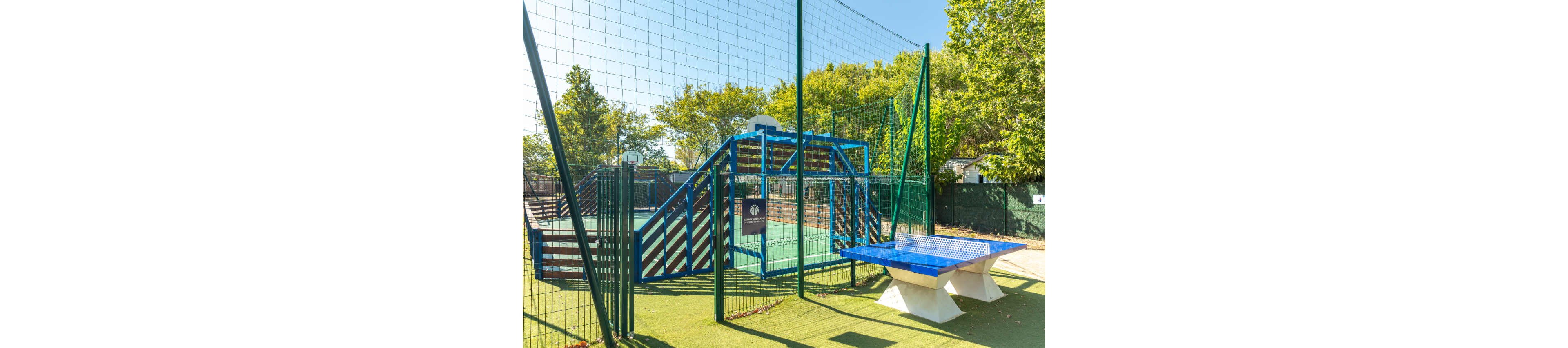 An outdoor sports court features a blue multi-level playground structure with stairs and railings. The area is enclosed by tall green metal fencing and surrounded by lush trees. A blue and white outdoor ping pong table stands in the foreground on artificial grass. Bright daylight and clear skies create a vibrant, recreational atmosphere.