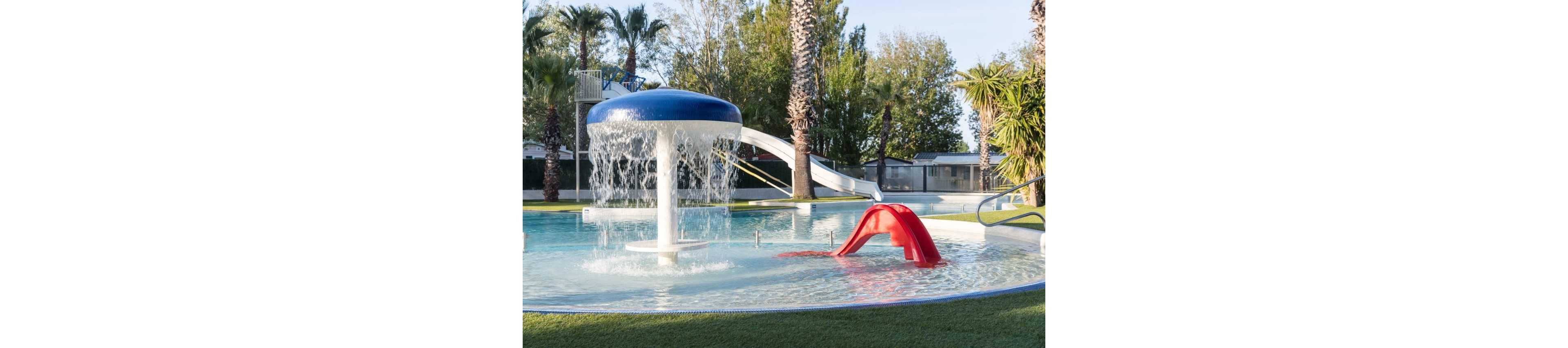 An outdoor splash pool features a large mushroom-shaped water fountain with cascading streams. A small red slide and a longer white slide curve into the shallow pool, suggesting a family-friendly water play area. Palm trees and other greenery surround the pool, creating a relaxed resort-like atmosphere. The scene is brightly lit, emphasizing clear blue water and playful structures.