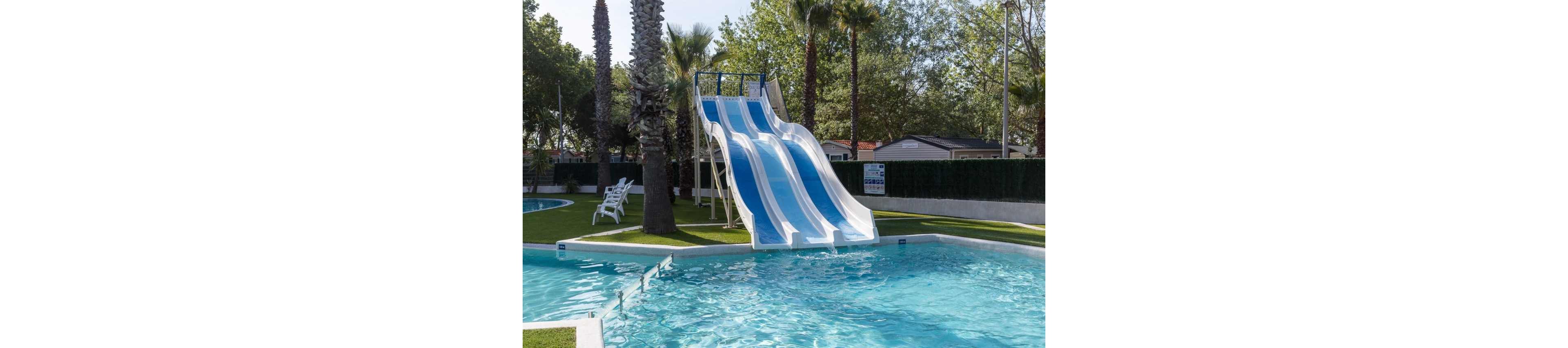 An outdoor swimming pool features a large triple-lane water slide with blue and white stripes. The slide descends into a clear blue pool surrounded by neatly trimmed grass and palm trees. White poolside chairs sit near the edge, and a hedge separates the pool area from nearby buildings. The scene suggests a quiet, sunny day at a family-friendly resort or campground.